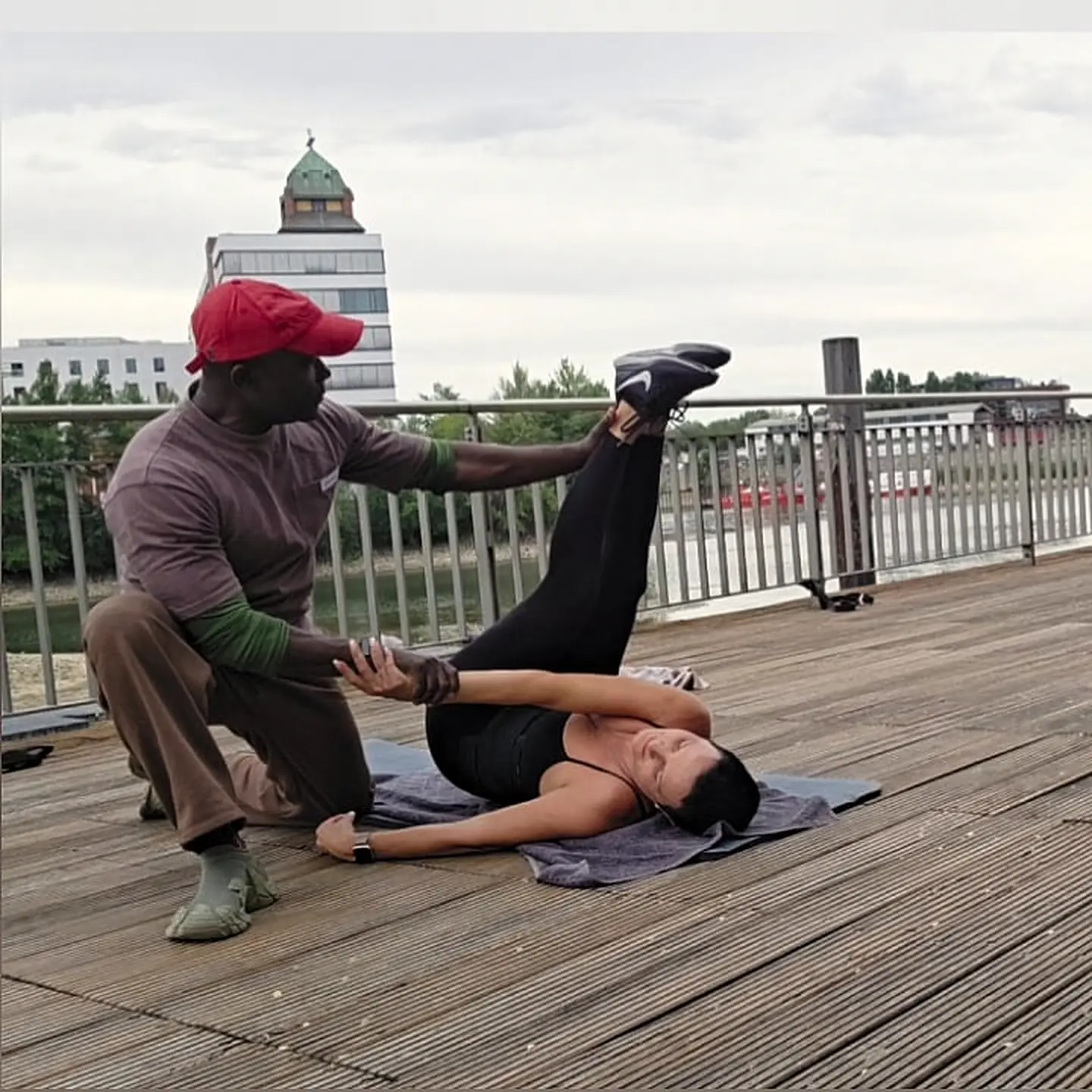 Trainer führt eine Dehnübung mit einer Kundin auf einem Holzsteg am Wasser